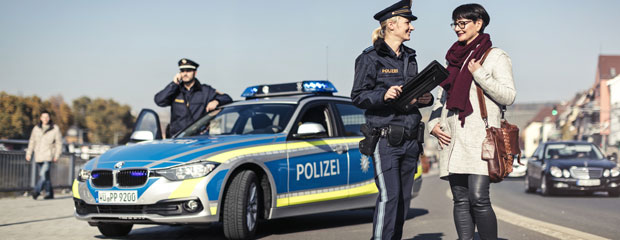 Polizei im Bürgerdialog Im Vordergrund ist eine Polizeibeamtin auf der Straße im Dialog mit einer Bürgerin zu sehen. Dahinter befindet sich ein uniformiertes Polizeifahrzeug, neben dem ein Polizeibeamter steht, der einen Funkspruch absetzt.
