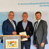 Innenminister Joachim Herrmann neben Alexander Brochier mit Bundesverdienstkreuz am Bande und Urkunde, rechts daneben eine weitere Person