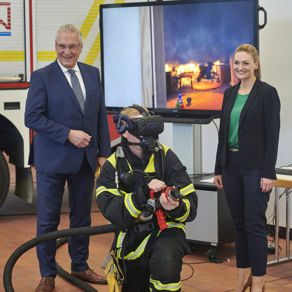 Herrmann und Gerlach neben Feuerwehrmann mit VR-Brille, dahinter Bildschirm mit Übungsszenario