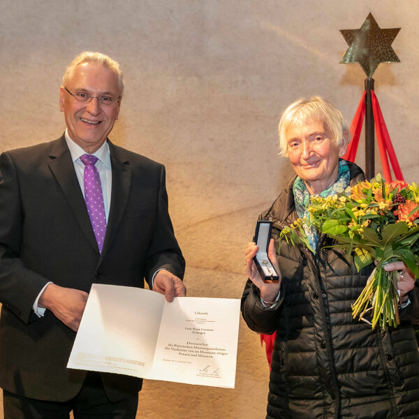 Innenminister Joachim Herrmann neben Rosa Carstens mit Blumenstrauß und Ehrenzeichen für ehrenamtliches kirchliches Engagement